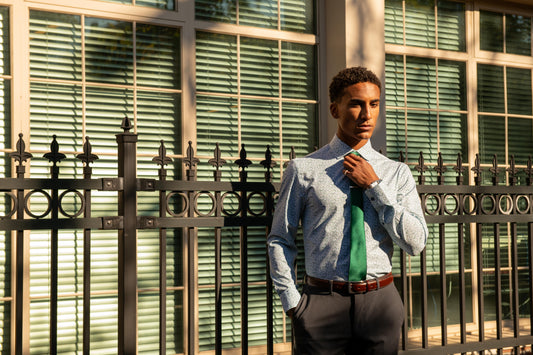 A man wearing a light blue patterned dress shirt, a green tie, and dark gray trousers stands in front of a window with green blinds and a black metal fence.