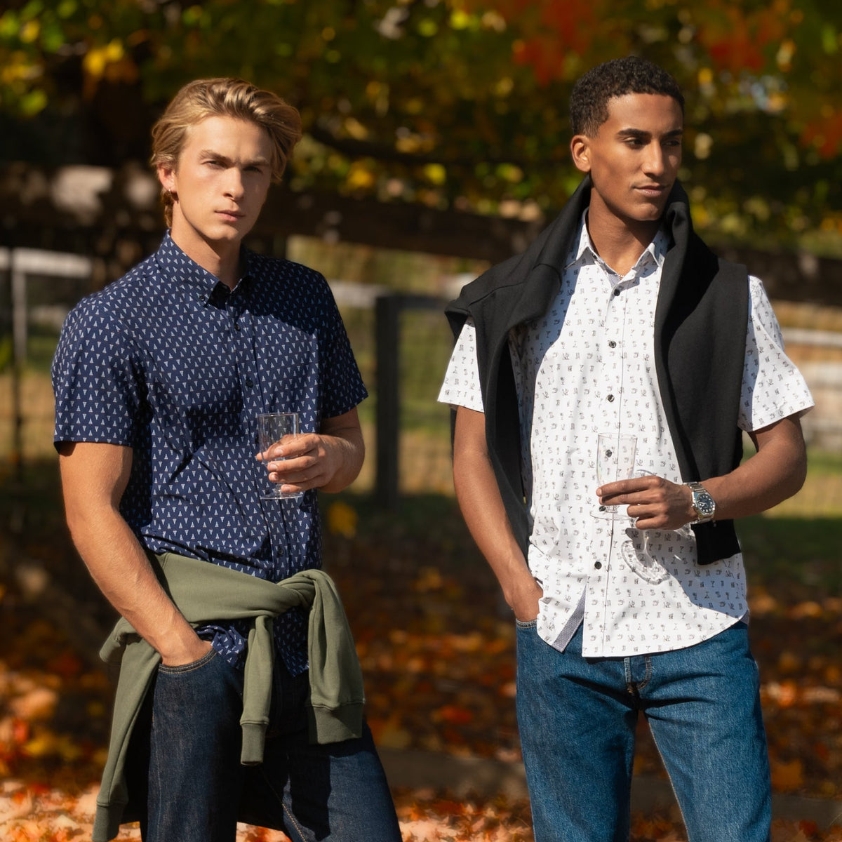 Two men standing outdoors in a farmyard covered with fallen autumn leaves, wearing casual shirts and jeans under a tree with orange and yellow leaves.
