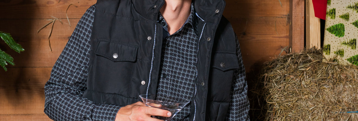 A man wearing a black quilted puffer vest over a dark blue and white checkered flannel shirt, standing in a rustic holiday-themed setting with a hay bale, wooden wall, and Christmas decorations.