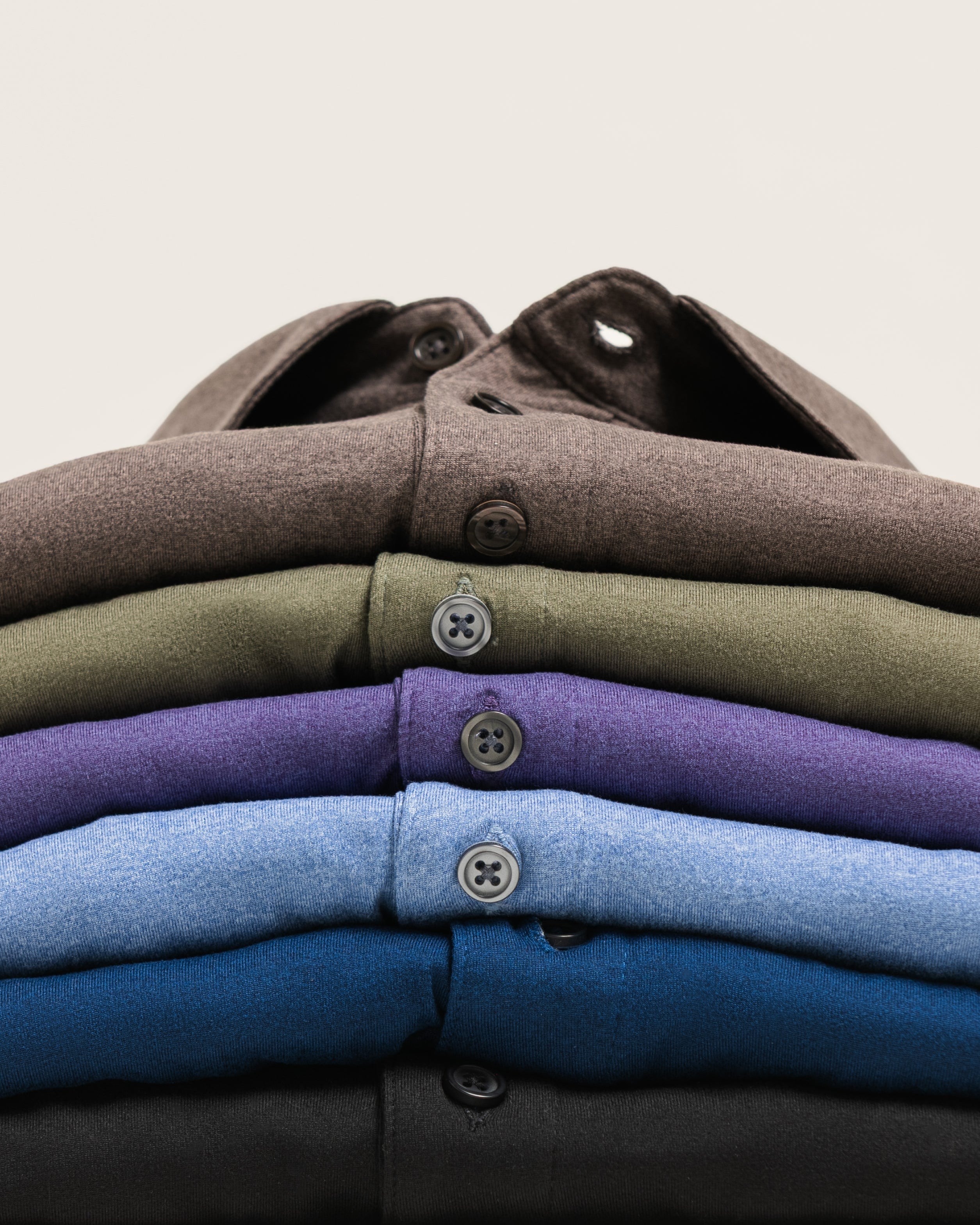 A stack of six folded button-up shirts in various colors, including brown, olive green, purple, light blue, navy, and black, arranged neatly on a light beige background.