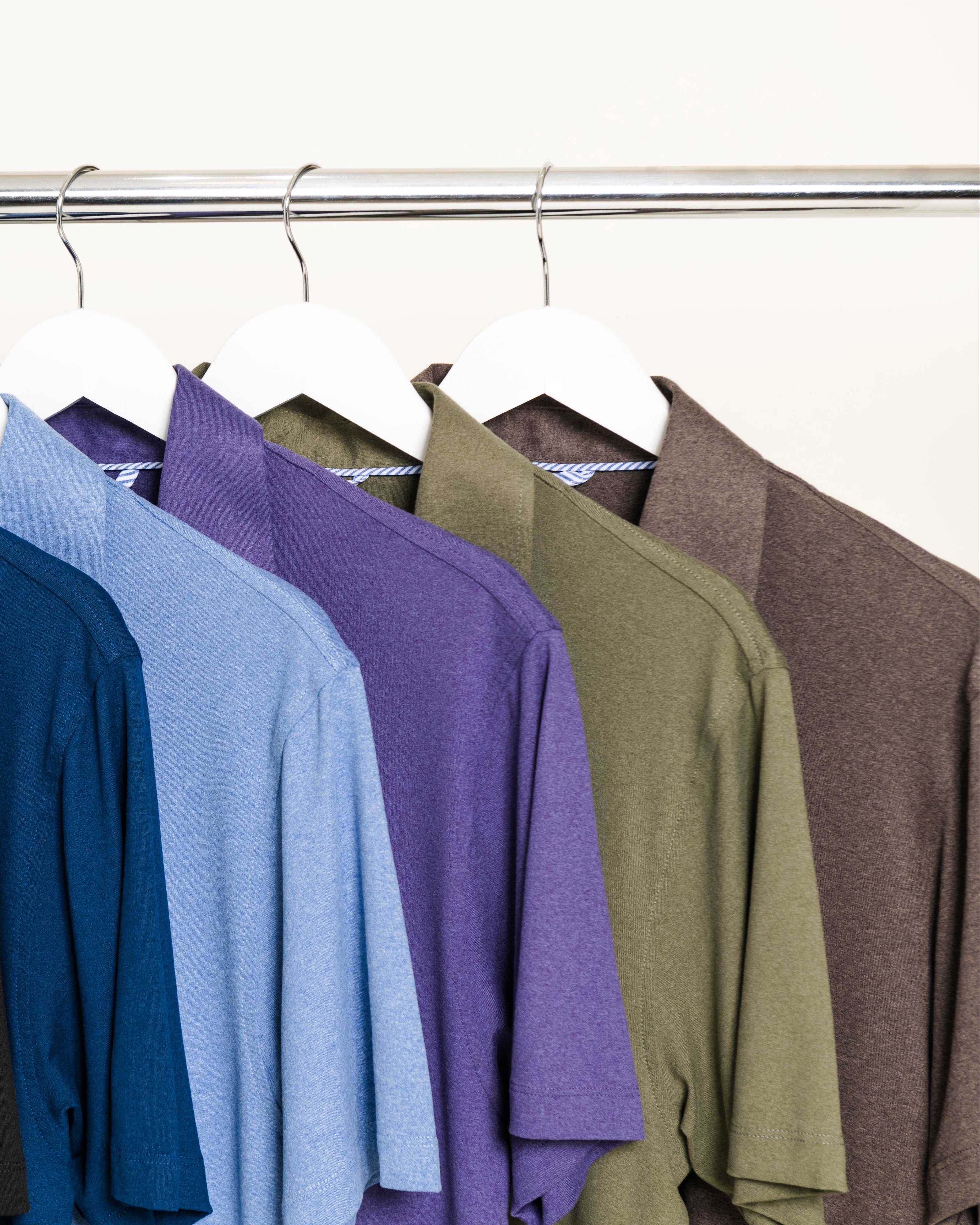 A row of men's short-sleeve polo shirts hanging on white hangers from a metal rack, shown in black, blue, light blue, purple, olive green, and brown.