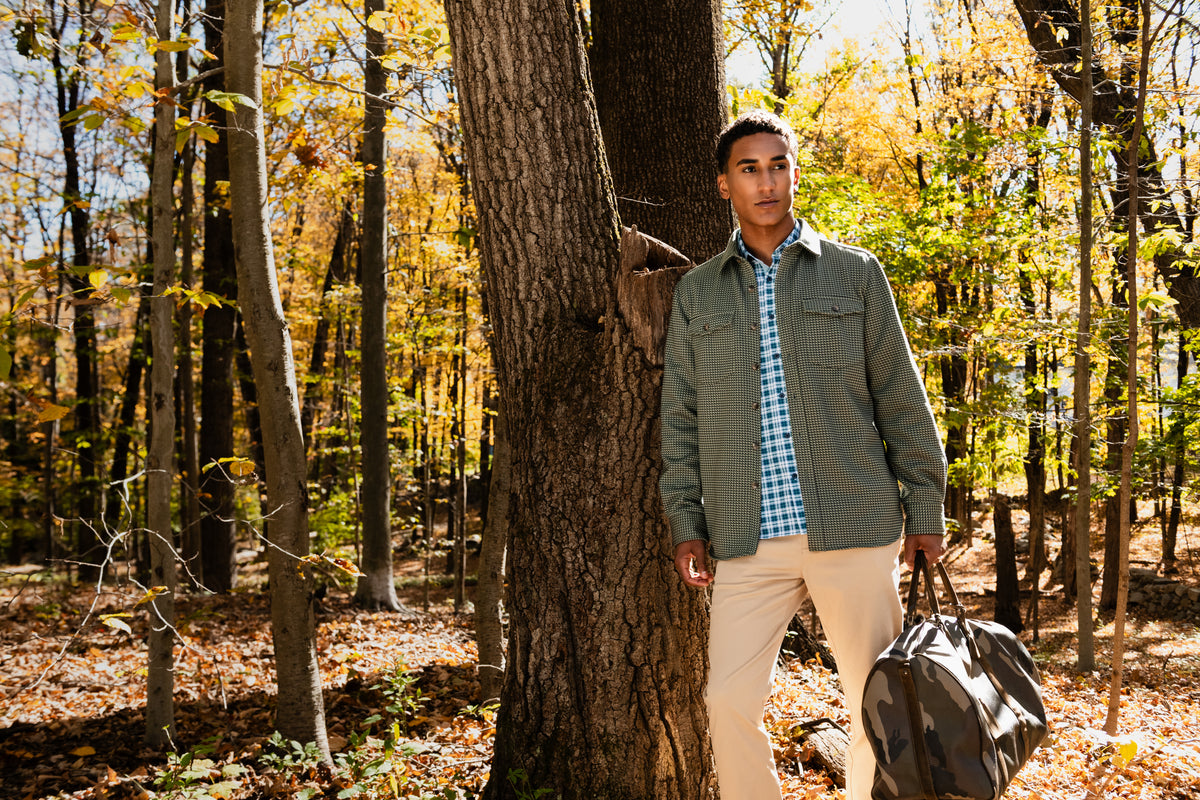 A man standing in a sunlit autumn forest next to a large tree trunk, wearing an olive green overshirt over a blue-and-white check shirt with light beige pants, and carrying a dark duffel bag.