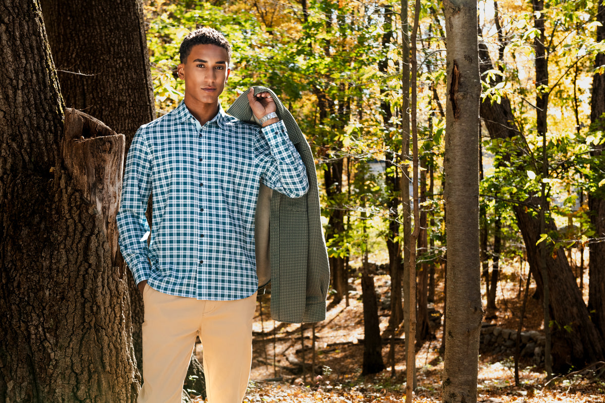 A man standing in a wooded forest setting, wearing a blue and white checkered long-sleeve button-down shirt, light beige pants, and holding a gray blazer over his shoulder.
