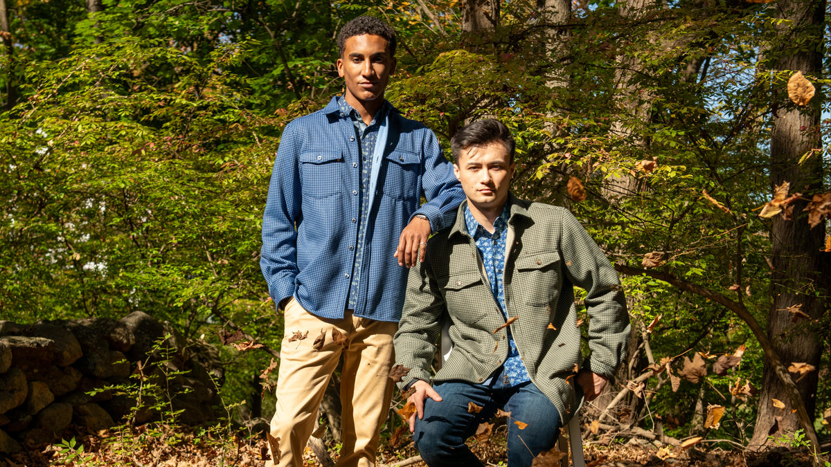 Two men posing outdoors in a wooded setting. The standing man wears a blue button-up shirt over a light blue collared shirt with beige pants, and the kneeling man wears an olive green jacket over a blue patterned shirt with dark jeans.