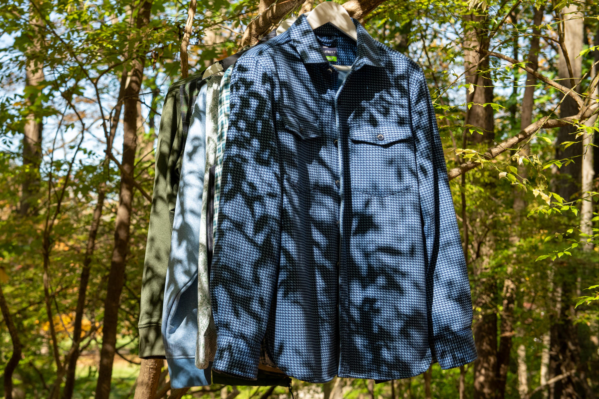 A blue patterned button-up shirt hangs on a wooden hanger from a tree branch in a sunlit forest, with two additional shirts partially visible behind it.