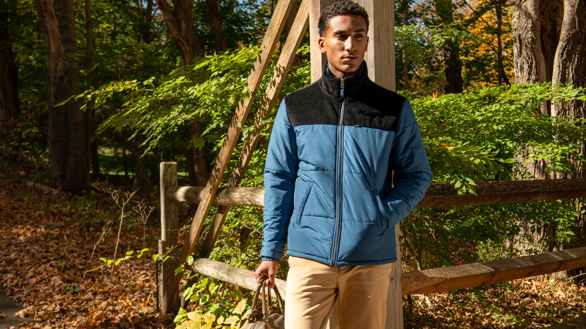 A man wearing a blue and black zip-up jacket and tan pants stands in a wooded outdoor setting next to a wooden fence, holding a gray duffel bag.