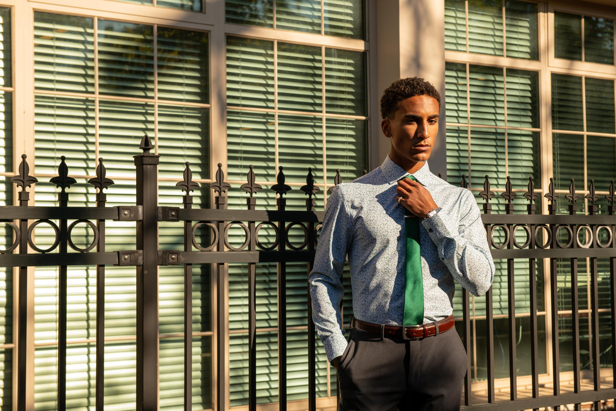 A man wearing a light blue patterned dress shirt, a green tie, and dark gray trousers stands in front of a window with green blinds and a black metal fence.