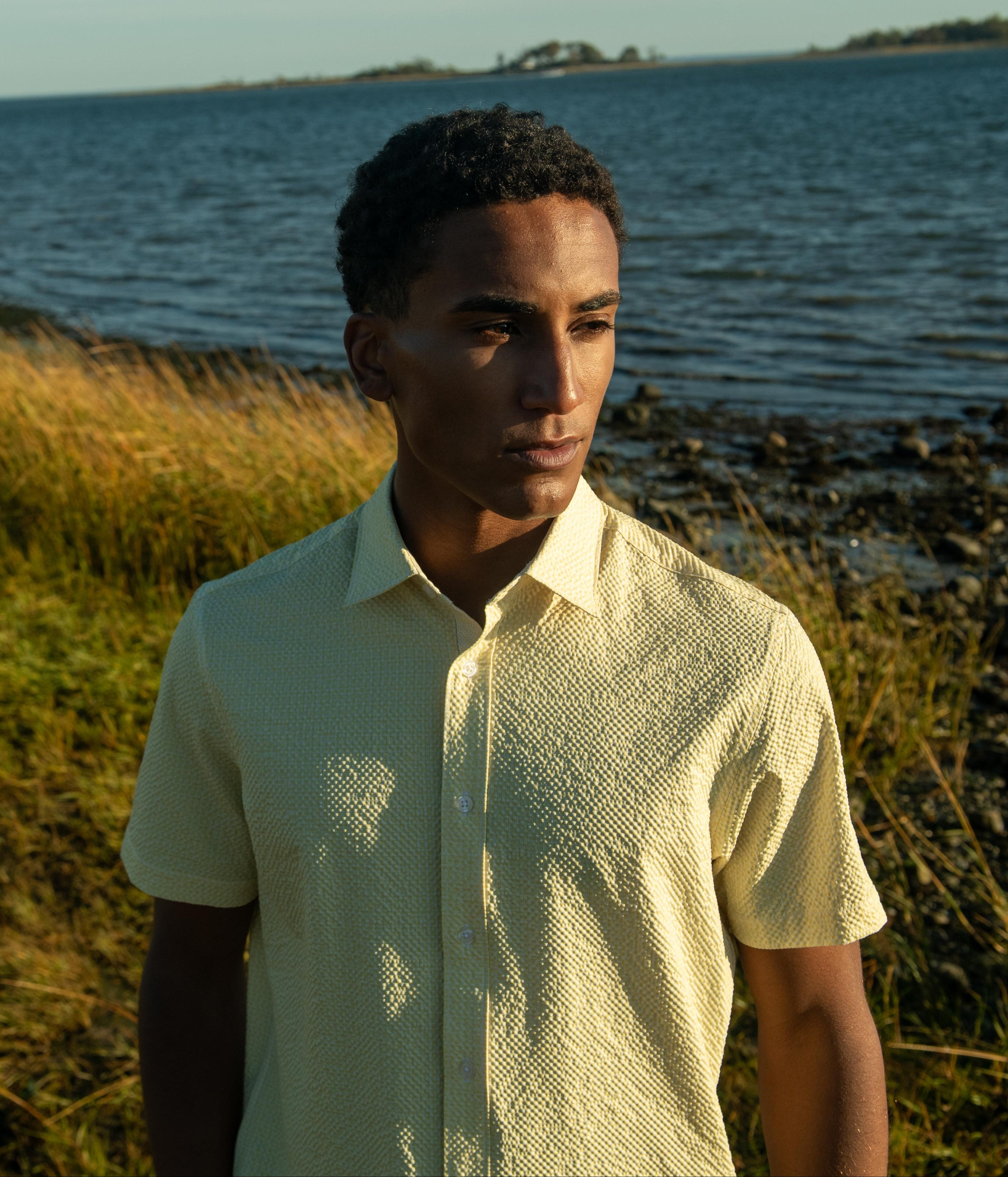 A man wearing a pale yellow textured short-sleeve button-up shirt stands outdoors on a grassy shoreline with water and distant land visible in the background.